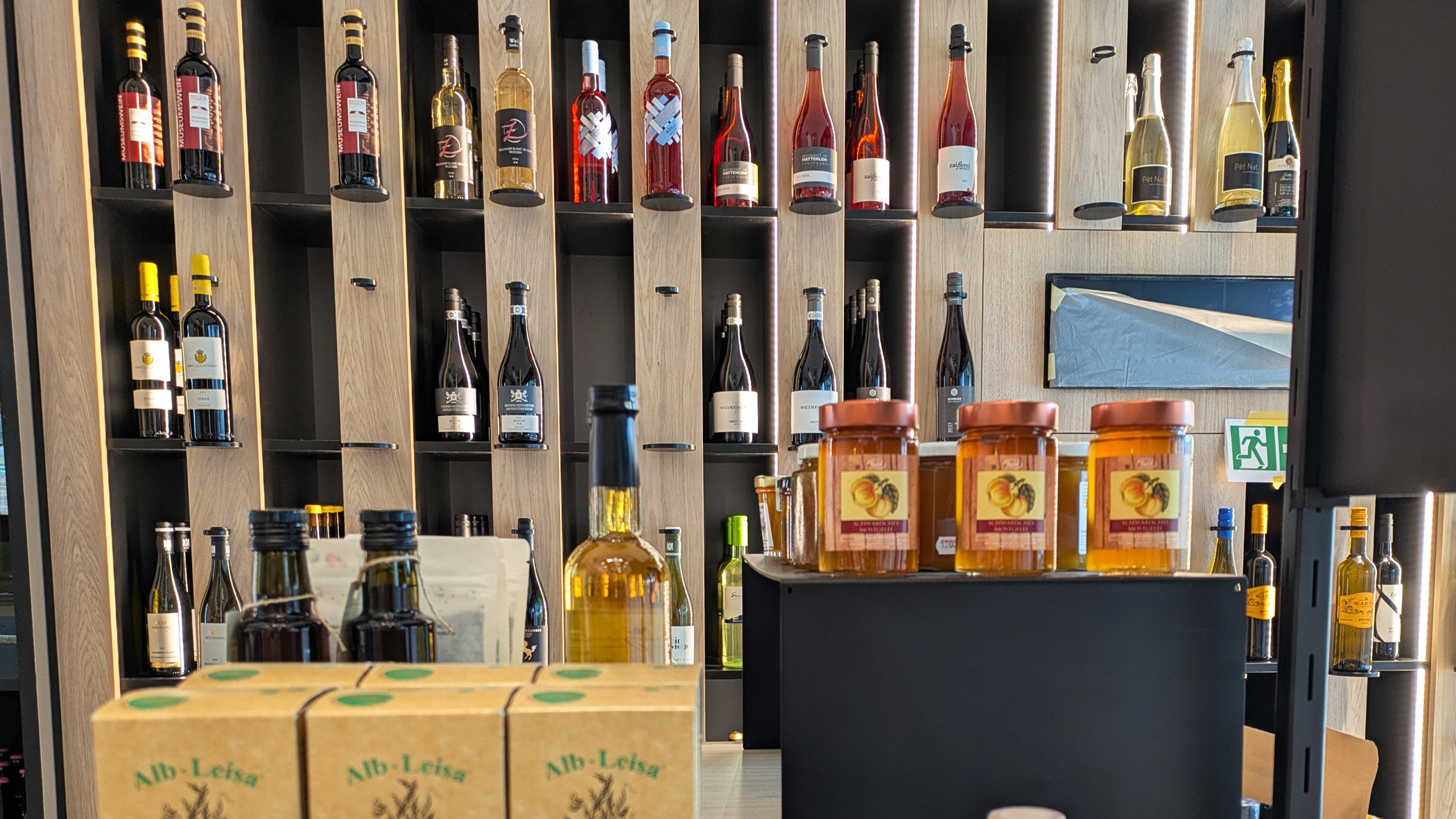 In einem Regal, dass eine gesamte Wandfläche einnimmt, stehen einzelne Flaschen unterschiedlicher Weine. Im Vodergrund sind andere Produkte, wie Alb-Linsen und Marmelade, ausgestellt.