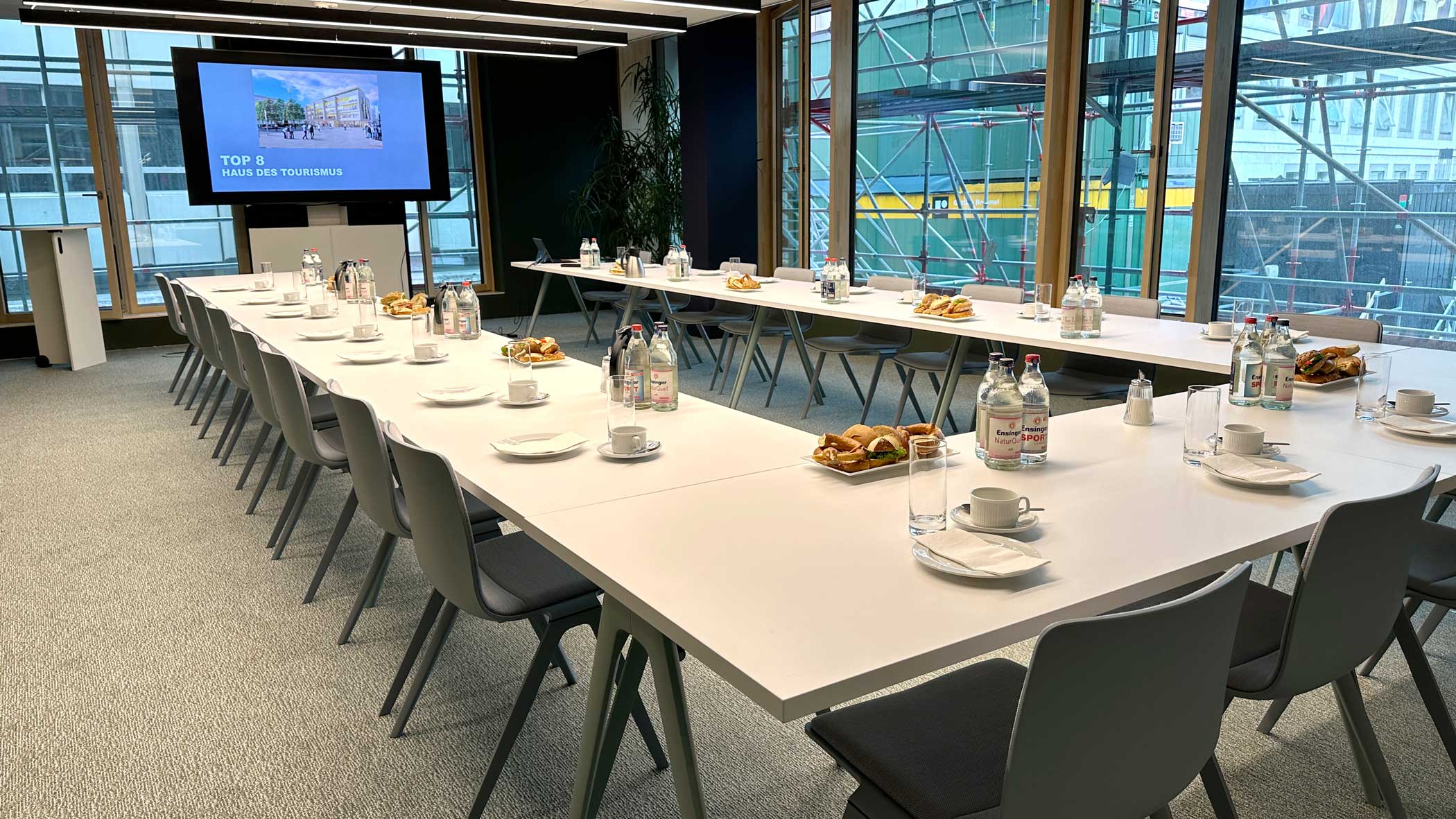 Besprechungszimmer E1, Tische mit Stühlen in U-Form in Richtung eines großen Bildschirms angeordnet. Jeder Platz ist eingedeckt, es stehen Kaffeekannen, Wasserflaschen und belegte Brote bereit.