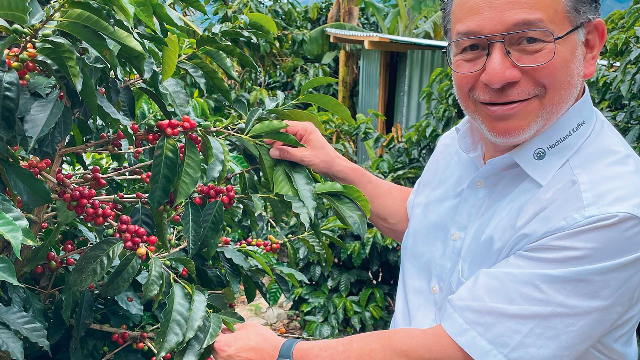 Rote Früchte an einer Kaffeepflanze. Ein freundlich schauender Mann mit Brille und weißem Hemd präsentiert die vollen Zweige der Pflanze.