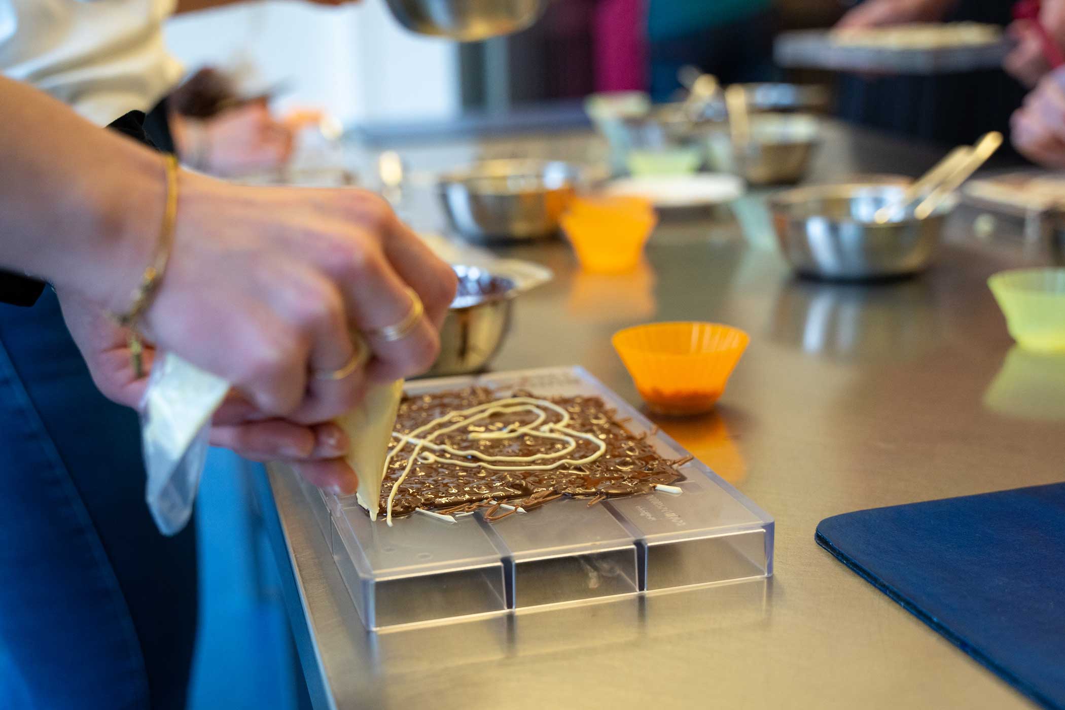 Eine Person verziert eine dünne Tafel glänzende Schokolade mit einer hellen Creme aus einer Spritztüte. Verschwommen im Hintergrund sind weitere Personen sowie Küchen-Utensilien zu sehen.