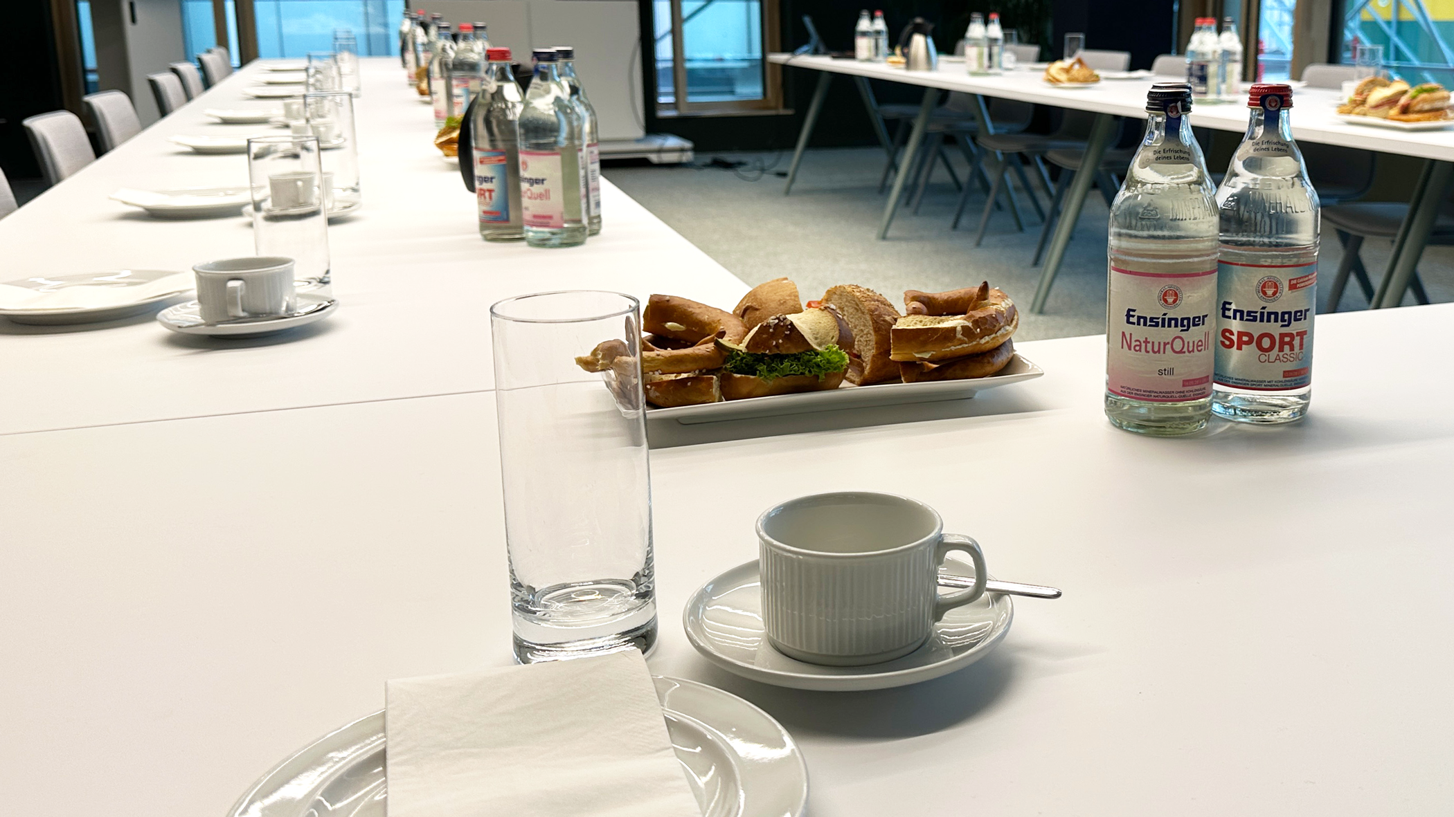 Auf den U-förmig angeordneten Tischen stehen an jedem Platz ein Teller, eine Tasse und ein Glas bereit. Ein paar Wasserflaschen stehen verteilt auf den Tischen, zudem ein paar belegte Brötchen und Butterbrezeln.