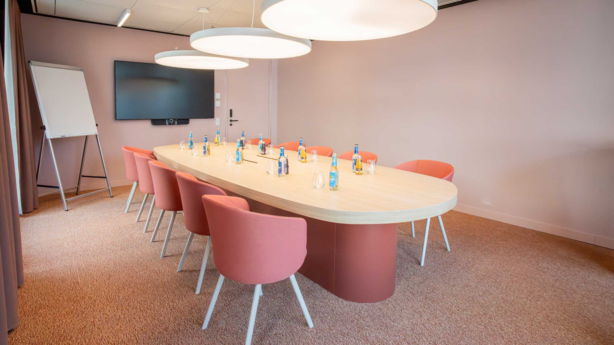 Rote Stühle angeordnet um einen ovalen Tisch; an der Stirnseite des Raums sind ein großer Bildschirm und ein Flipchart.