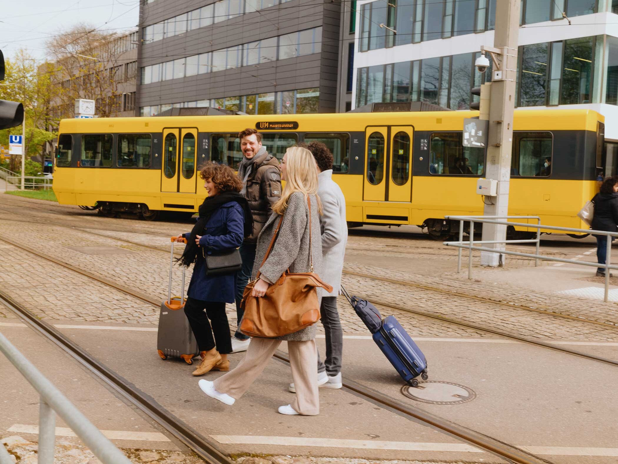 Eine Gruppe junger Leute mit Gepäck überquert eine Straße, im Hintergrund durchquert eine gelbe SSB U-Bahn das Bild.