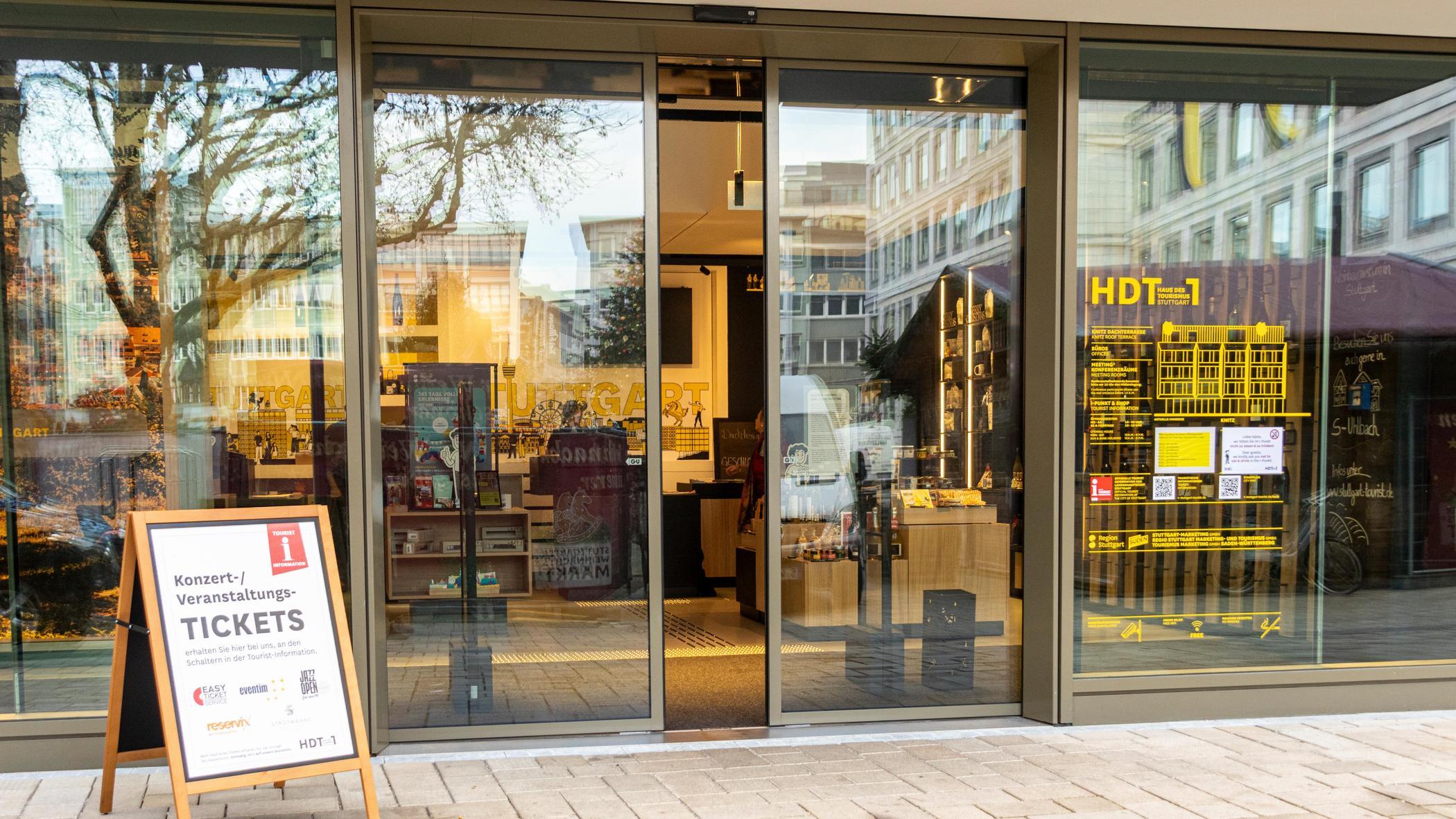 Eingang des Hauses des Tourismus in Stuttgart mit automatischen Glas-Schiebetüren. Ein Schild informiert über Veranstaltungstickets. Moderne und einladende Atmosphäre.