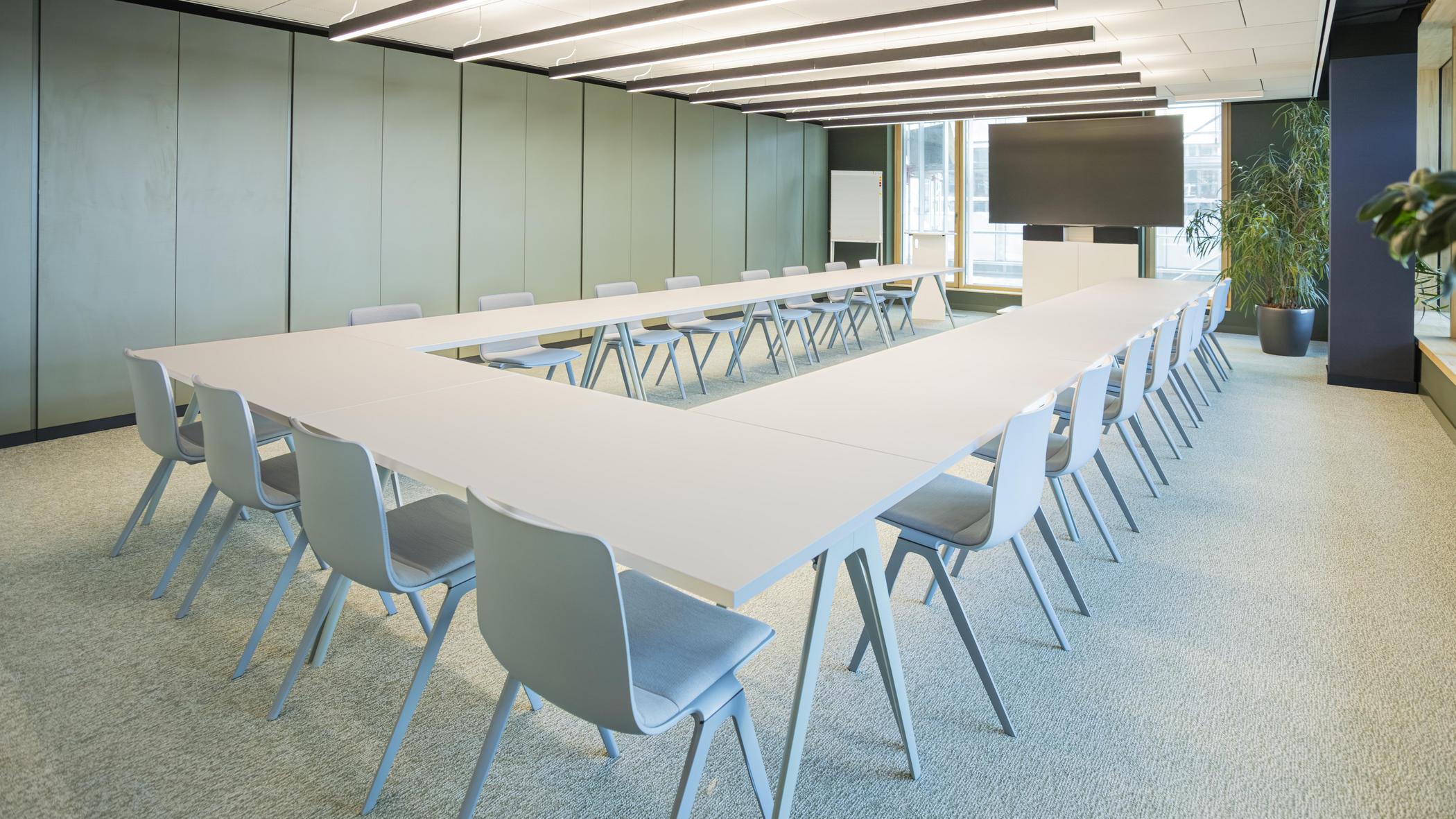 Tagungsraum Erlebnis, Klassischer Konferenzraum in gedeckten Farben mit U-förmig gestellten weißen Tischen und Bestuhlung. An der Stirnseite befindet sich ein großes Bildschirm.