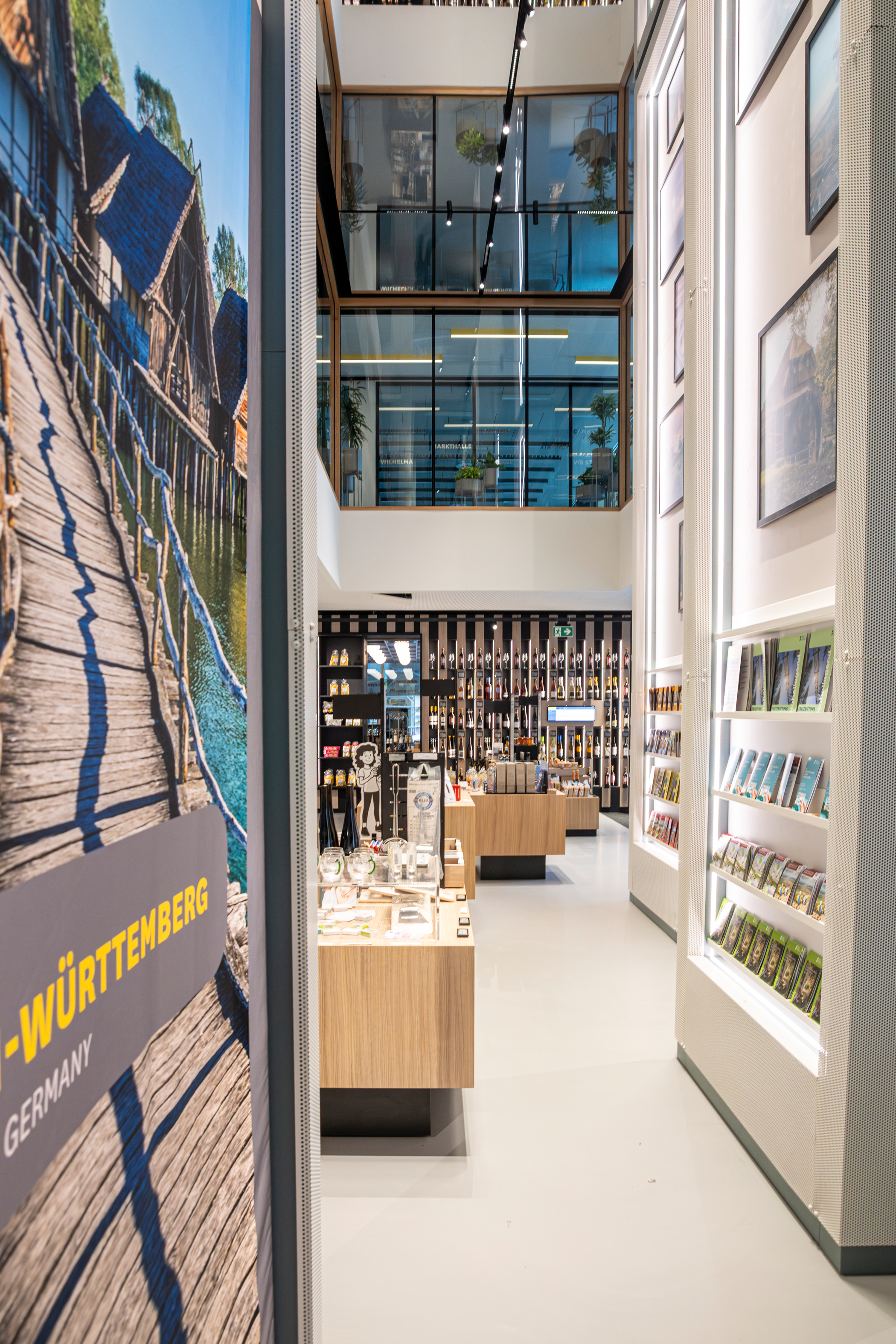 Blick in die Tourist-Information, Richtung Wein-Wand. Auslage von Produkten und Broschüren auf Tischen und in Regalen. Durch die hohe Decke ist ein Blick in die Ebene 1, wo sich die Tagungsräume befinden, möglich.