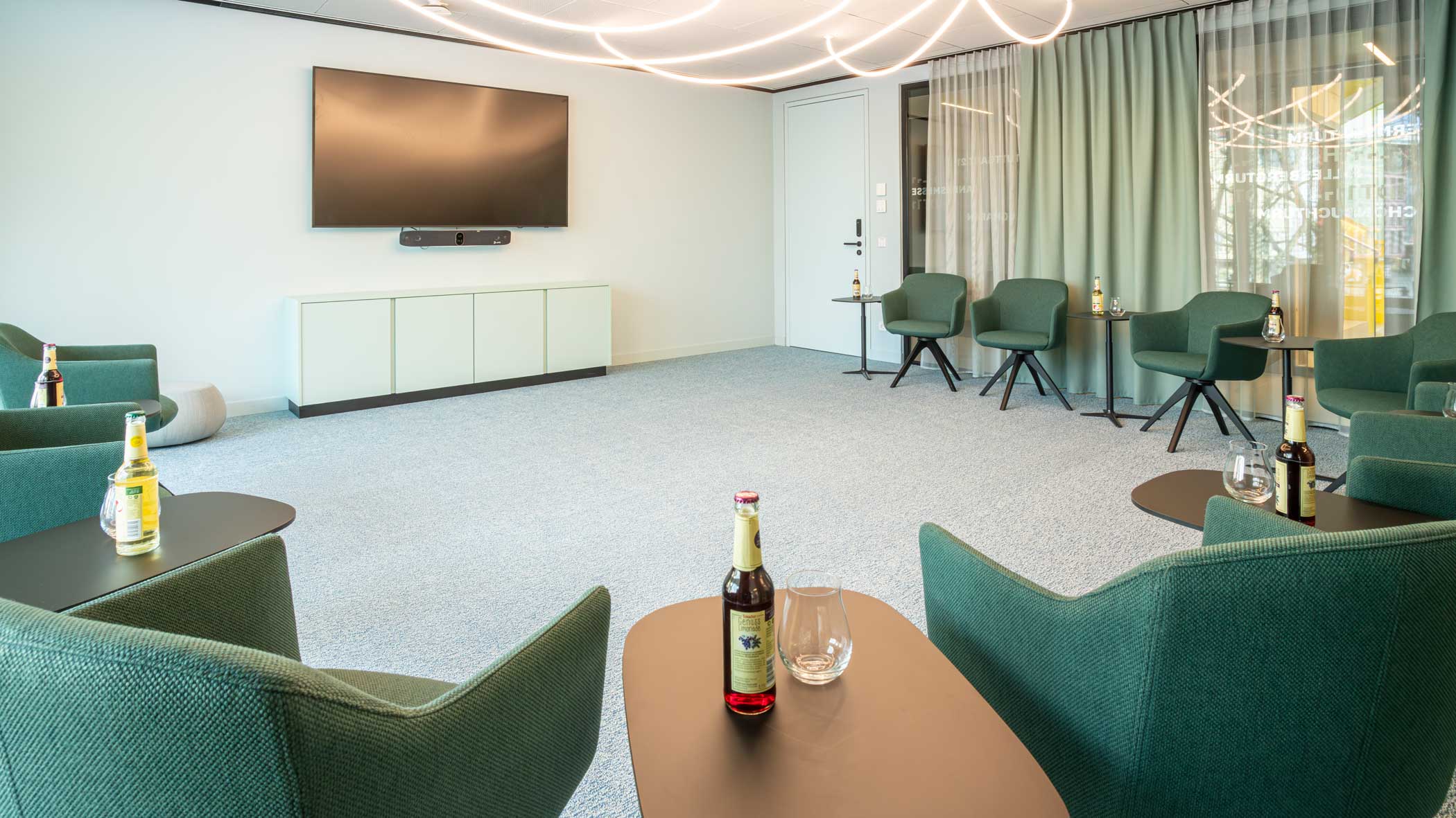 Grüne Stühle, die im Halbkreis angeordnet sind, mit je einem Beistelltisch, an der Stirnseite des Raums hängt ein flacher Bildschirm.