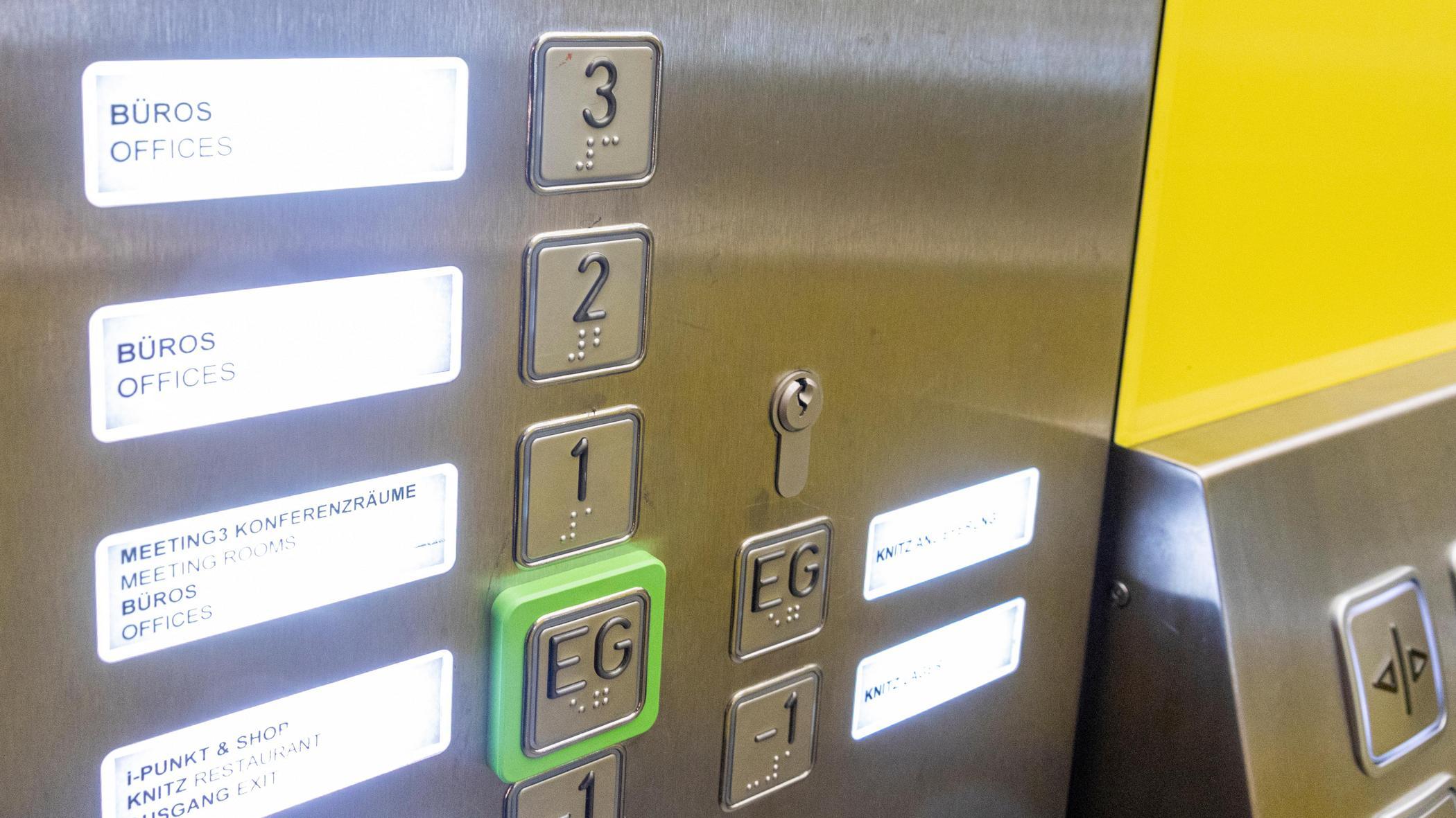 Ein Aufzugspanel mit Tasten in Brailleschrift und Beschriftungen in Deutsch und Englisch für Büros und Konferenzräume. Der Knopf für das Erdgeschoss ist grün hervorgehoben.