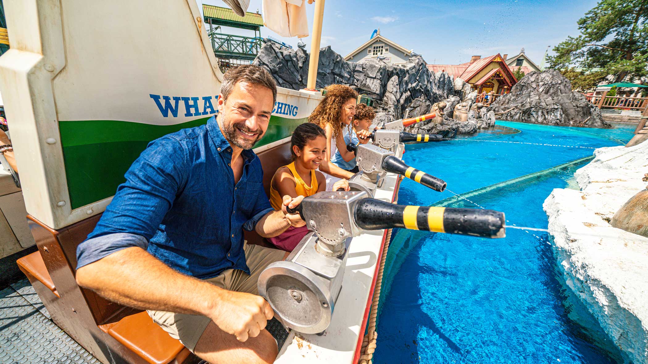 Ein Mann, eine Frau und zwei Kinder sitzen lachend in einer Art Boot auf Schienen. Sie spritzen mit großen an der Reling befestigten Spritzpistolen.