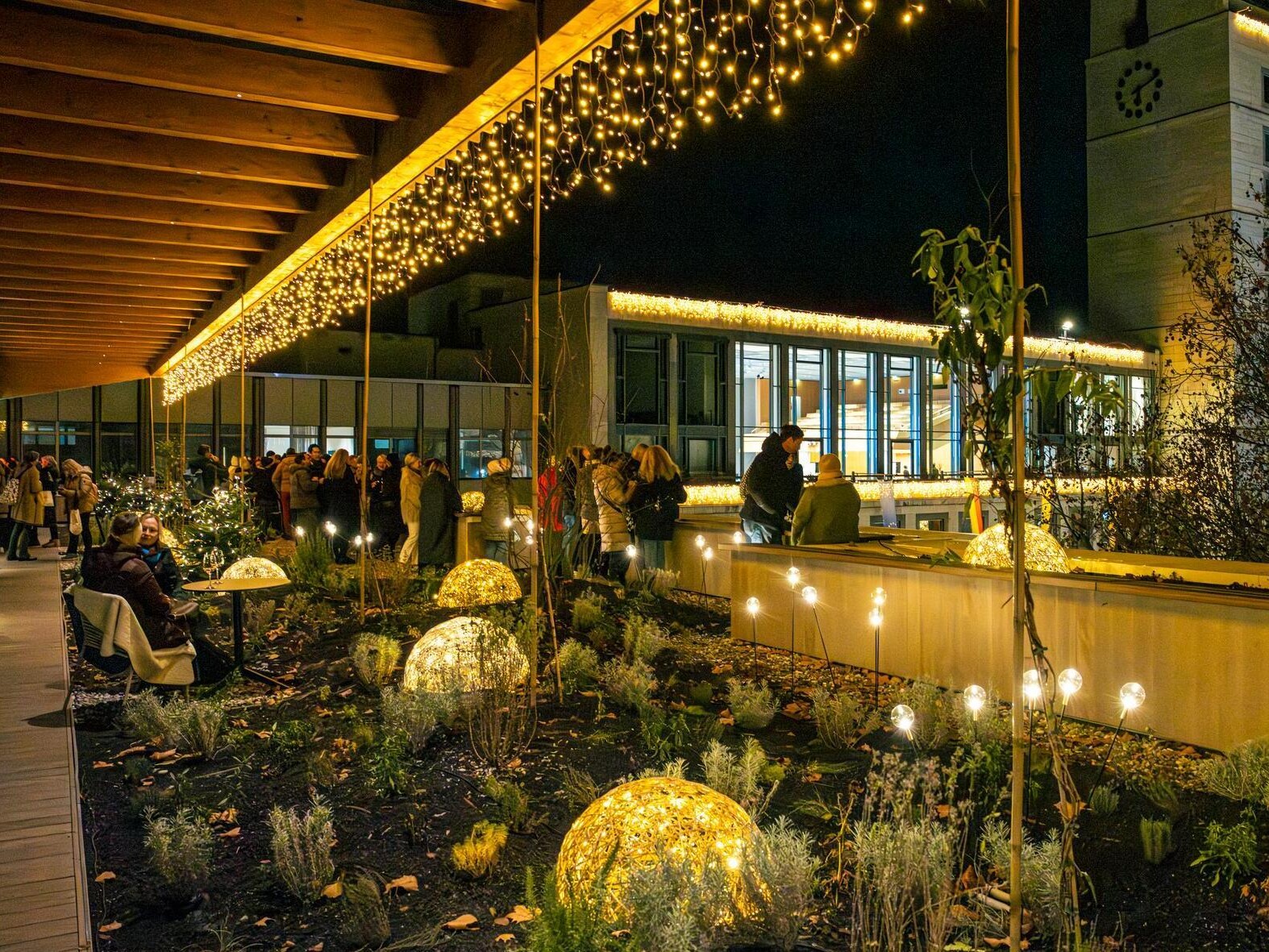 Dachterrasse des Haus des Tourismus mit vielen Lichtern: beleuchtete geflochtene Halbkugeln im Blumenbeet, Lichterkette am Dach und Leuchtkugeln auf Stäben. Im Hintergrund stehen Personen in warmer Kleidung zusammen.